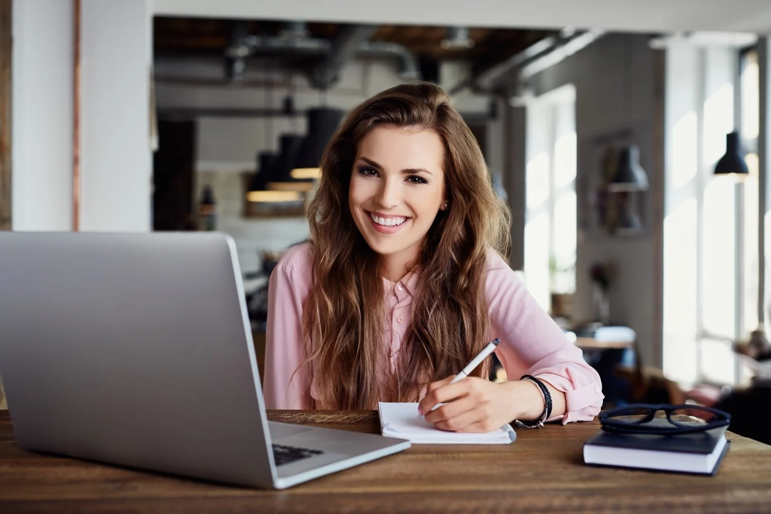 A smiling woman sitting at a desk, in front of a laptop, writing on a notepad.