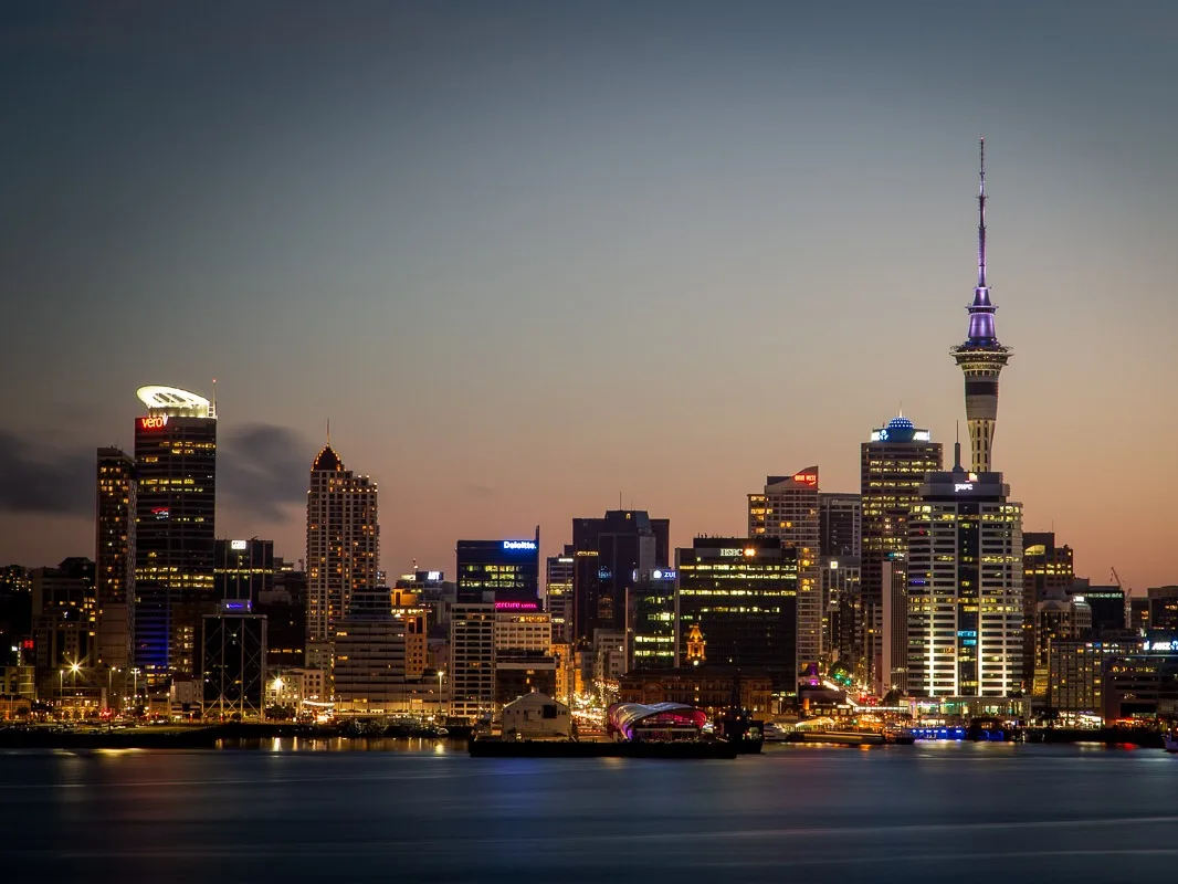 Auckland skyline at dusk with illuminated Sky Tower and waterfront buildings reflecting in harbor waters.