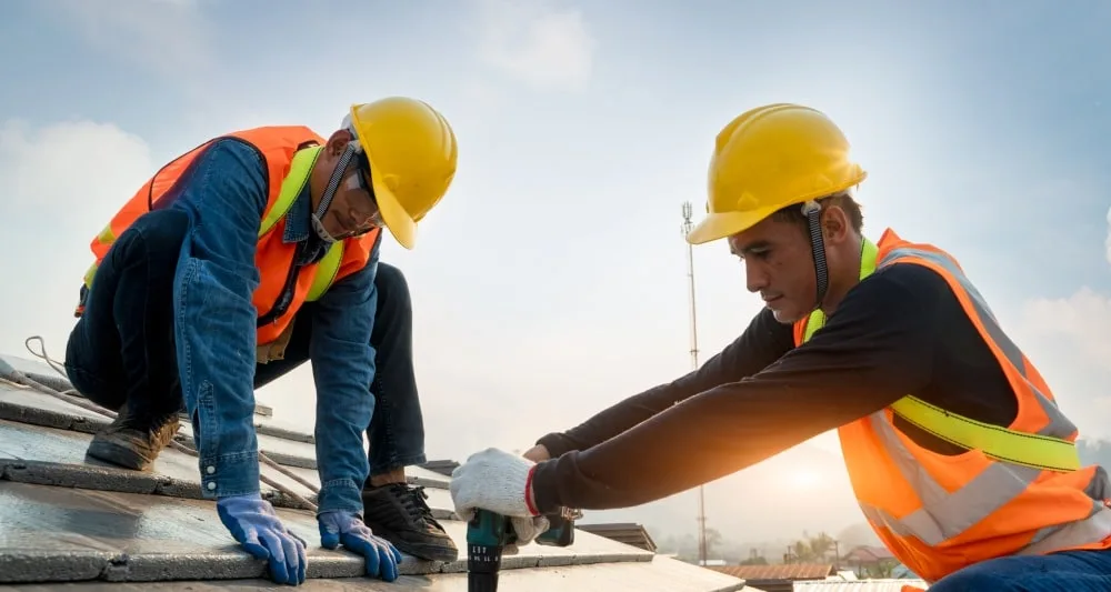 Two tilers working on a roof in protective hard hats and vests. 