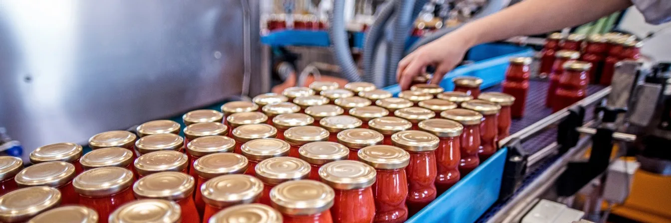 Worker inspecting rows of sealed red jars with gold lids on a blue production line in a food processing facility.