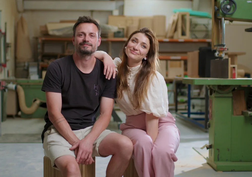 Husband and wife co-owners of furniture design business, Hegi Design House, sitting together in their warehouse surrounded by shelves stocked with their products.