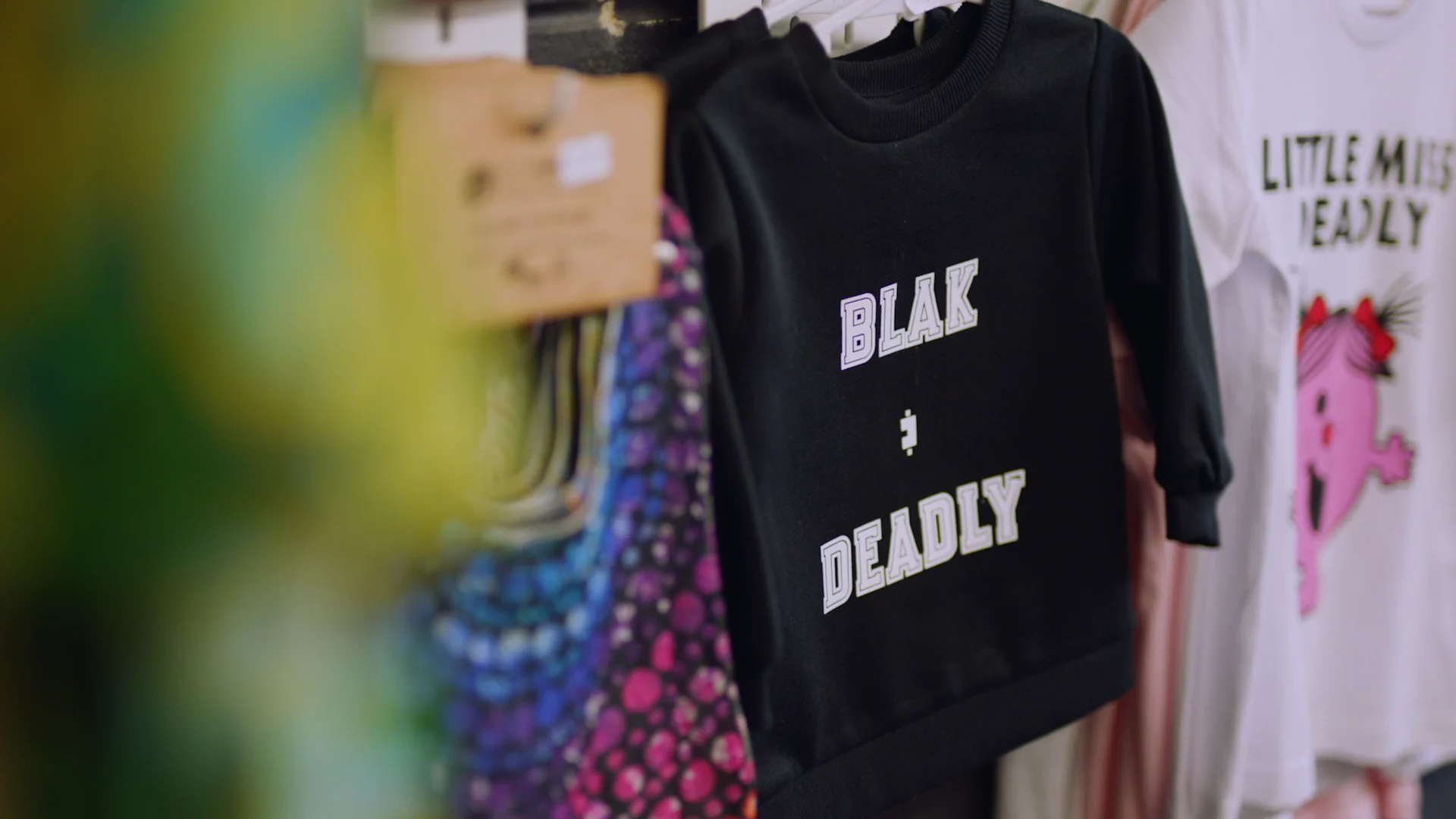 Black t-shirt with "BLAK + DEADLY" text hanging on rack next to white "LITTLE MISS DEADLY" shirt with pink character.
