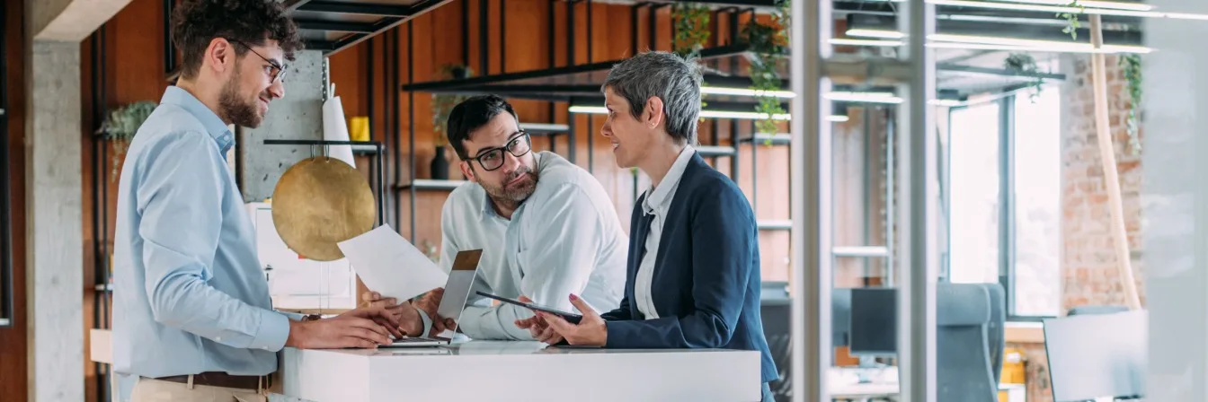 Three professionals in discussion around a white table in a modern office with wooden accents and industrial shelving.