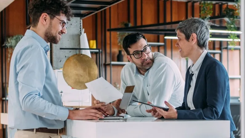 Three professionals in discussion around a white table in a modern office with wooden accents and industrial shelving.