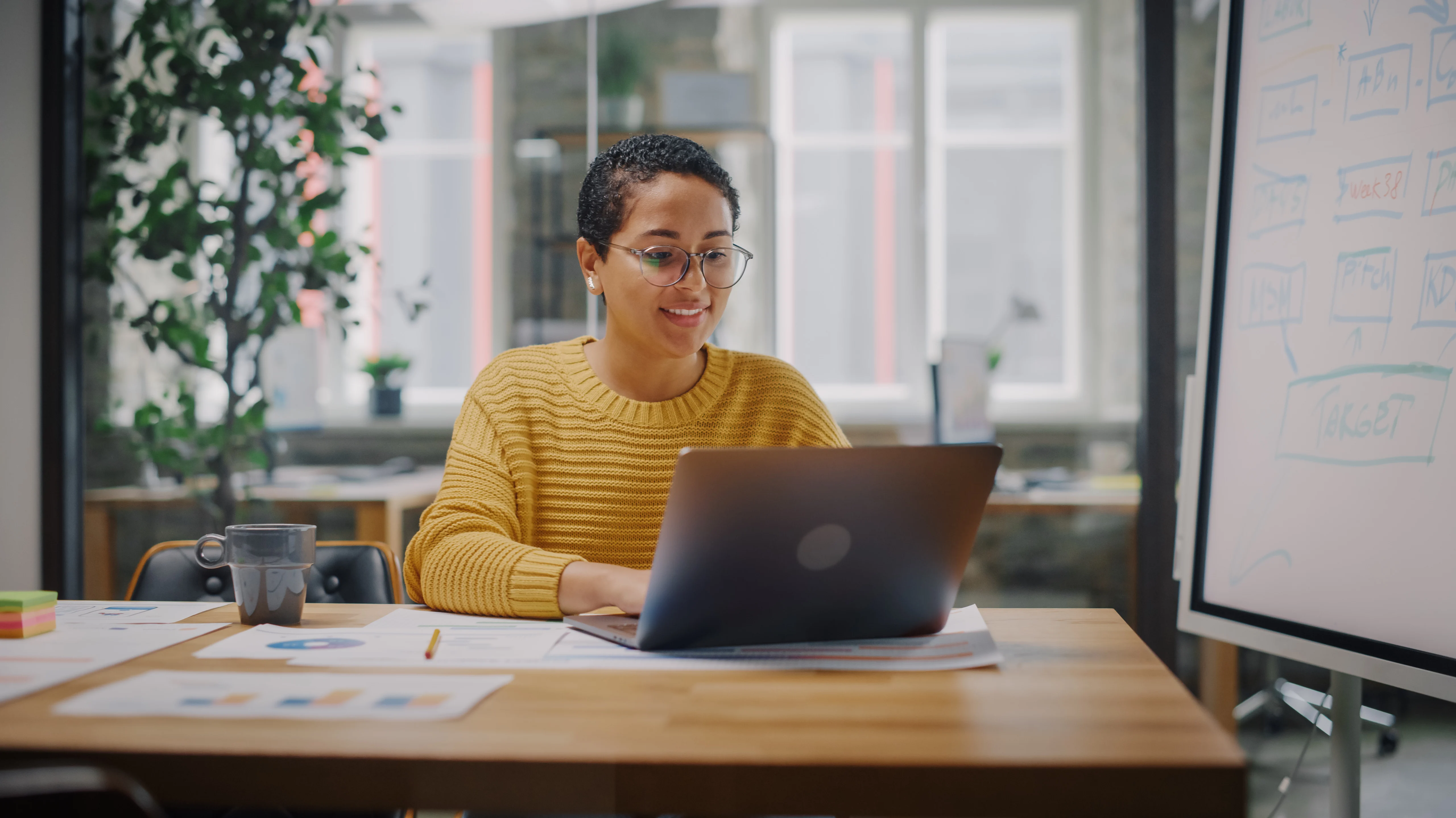 A young woman of colour sits at a desk in a bright yellow knitted jumper drinking a cup of coffee. She smiles while looking at a laptop. Nearby, a workflow is mapped out on a whiteboard.