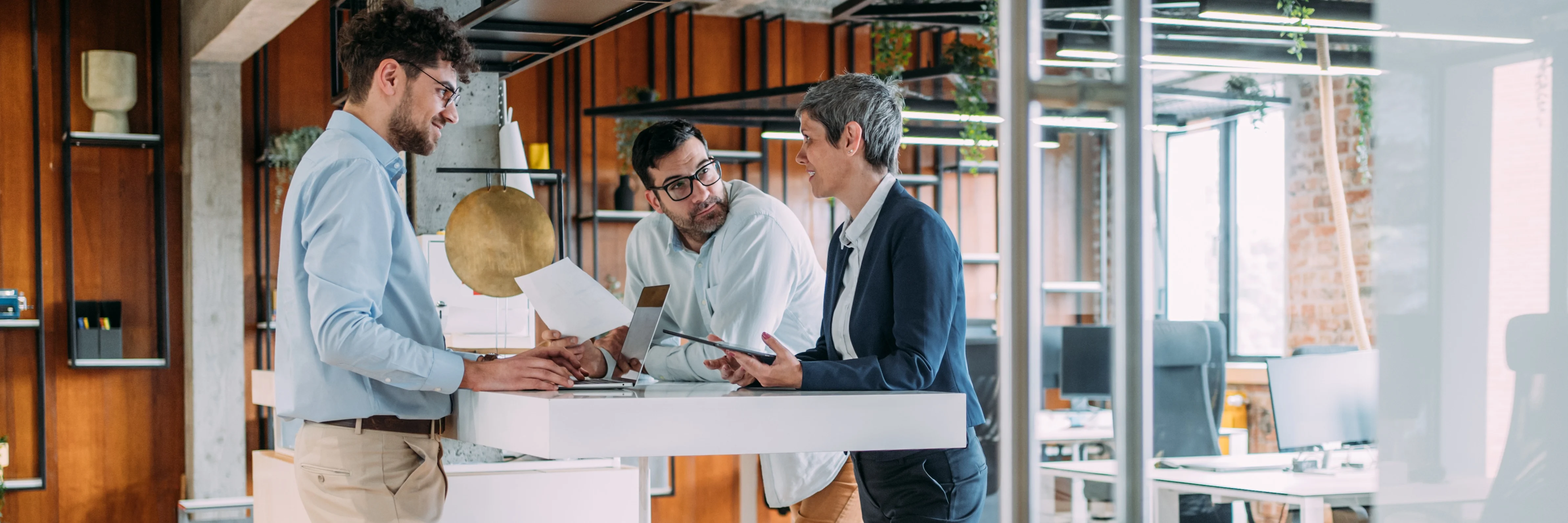 Three professionals discussing documents around a white table in a modern office with wooden walls and industrial design.
