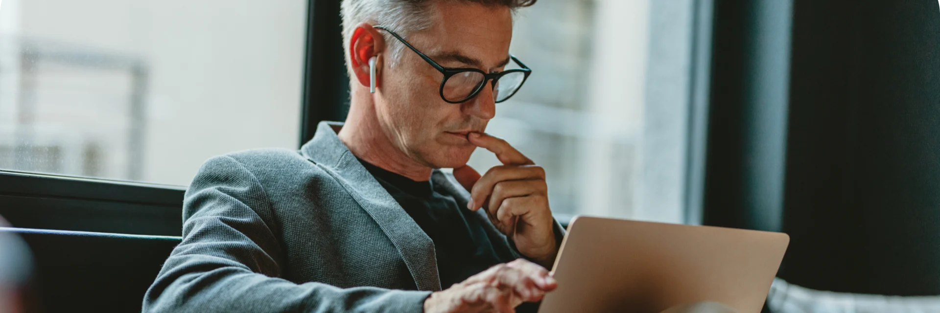 Professional in gray blazer working thoughtfully on laptop while wearing wireless earbuds.