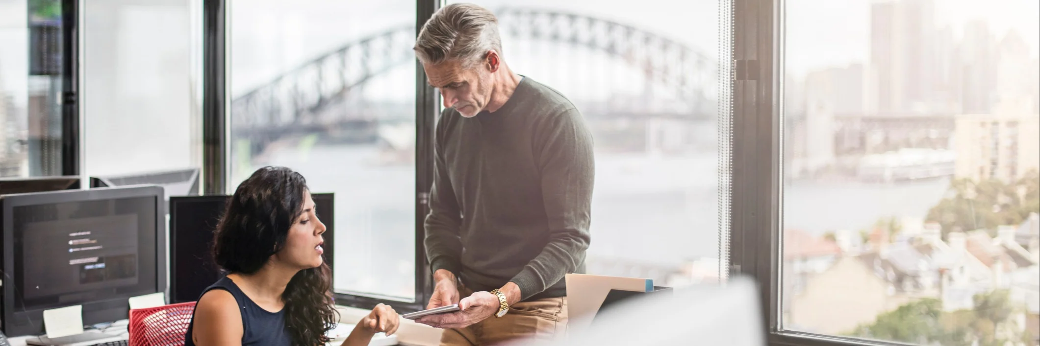 Two colleagues discussing work in a modern office with large windows overlooking Sydney Harbour Bridge.