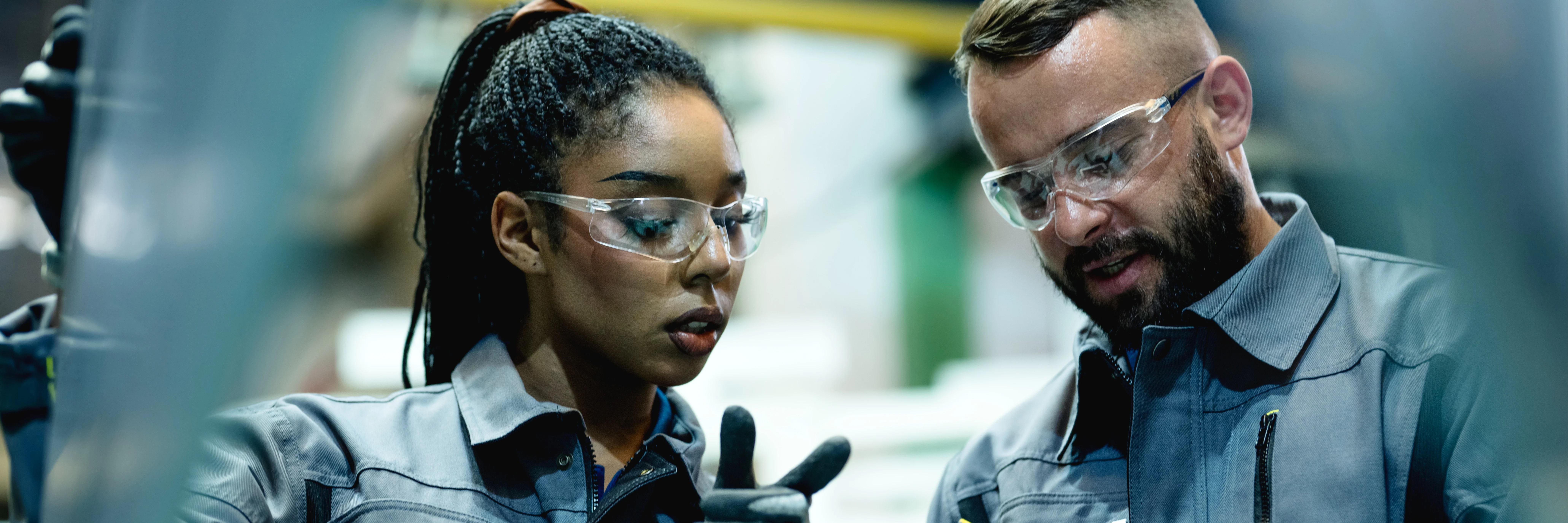 Two factory workers wearing safety glasses and gray uniforms discuss something on a digital tablet inside a manufacturing facility.