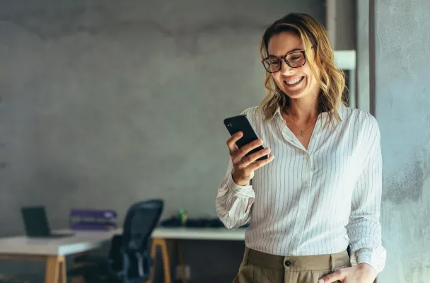 Smiling person standing in an office whilst looking at their mobile phone and smiling.