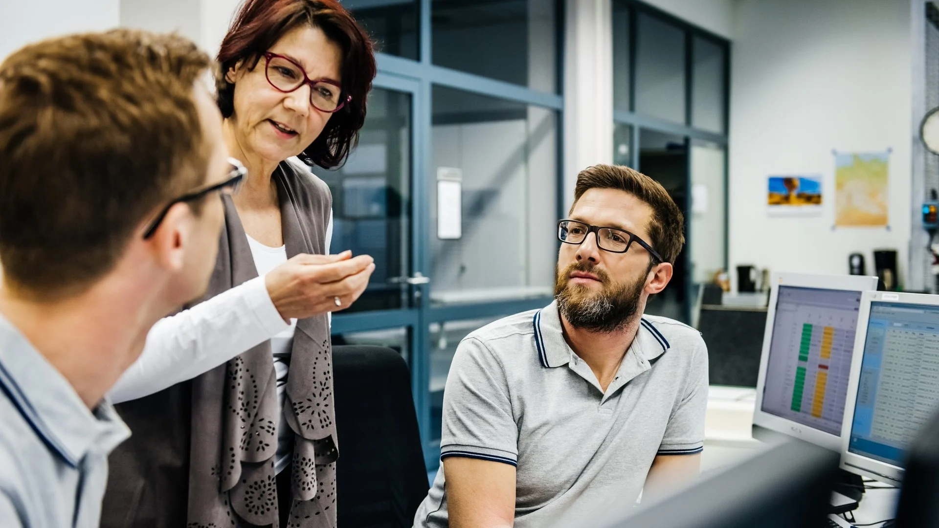 Three colleagues in discussion at an office workspace with computer monitors displaying data charts in background.