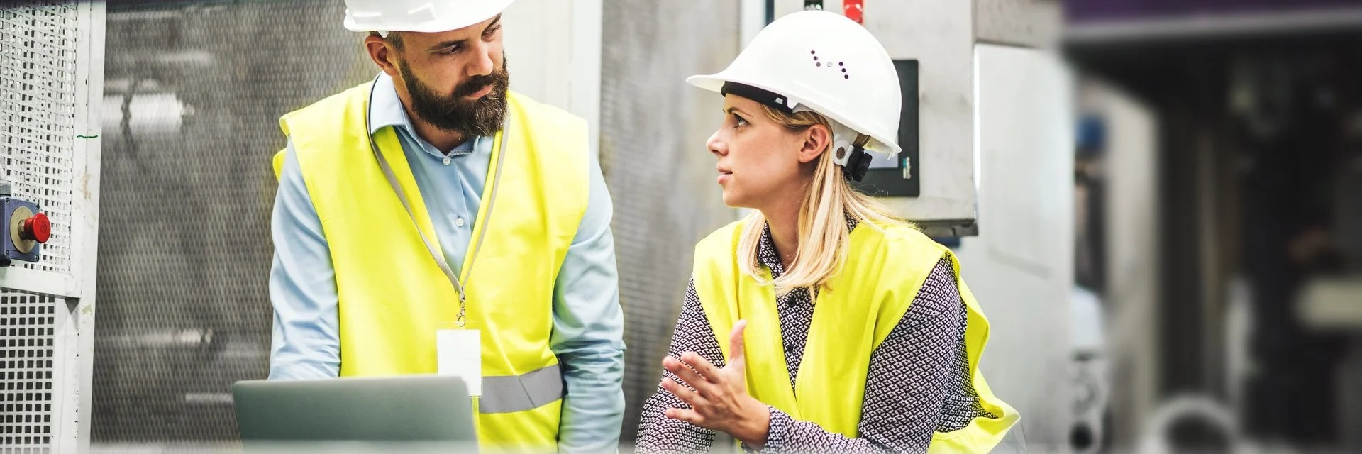 Two construction professionals in white hard hats and yellow safety vests discussing a project in an industrial setting.