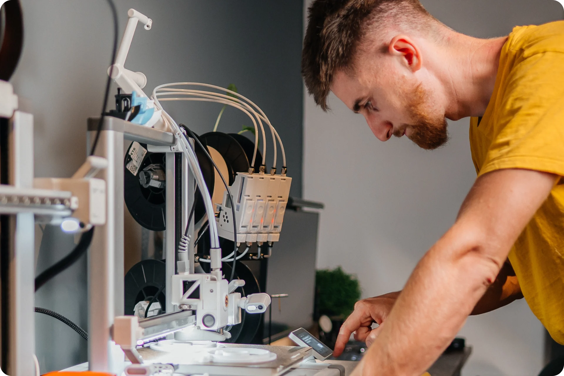 Person in yellow shirt examining a 3D printer with filament spools and electronic components in a workshop setting.