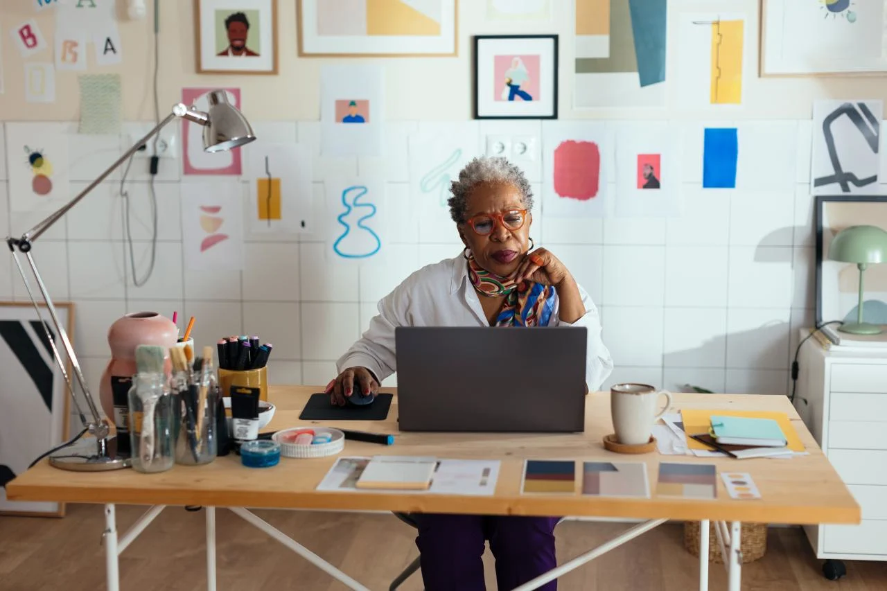 Focused senior woman with gray hair and trendy outfit sitting at table working on laptop in modern creative space full of artworks