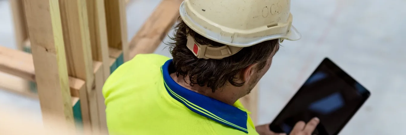 Construction worker in white hard hat and high-visibility yellow shirt using a tablet at a building site.