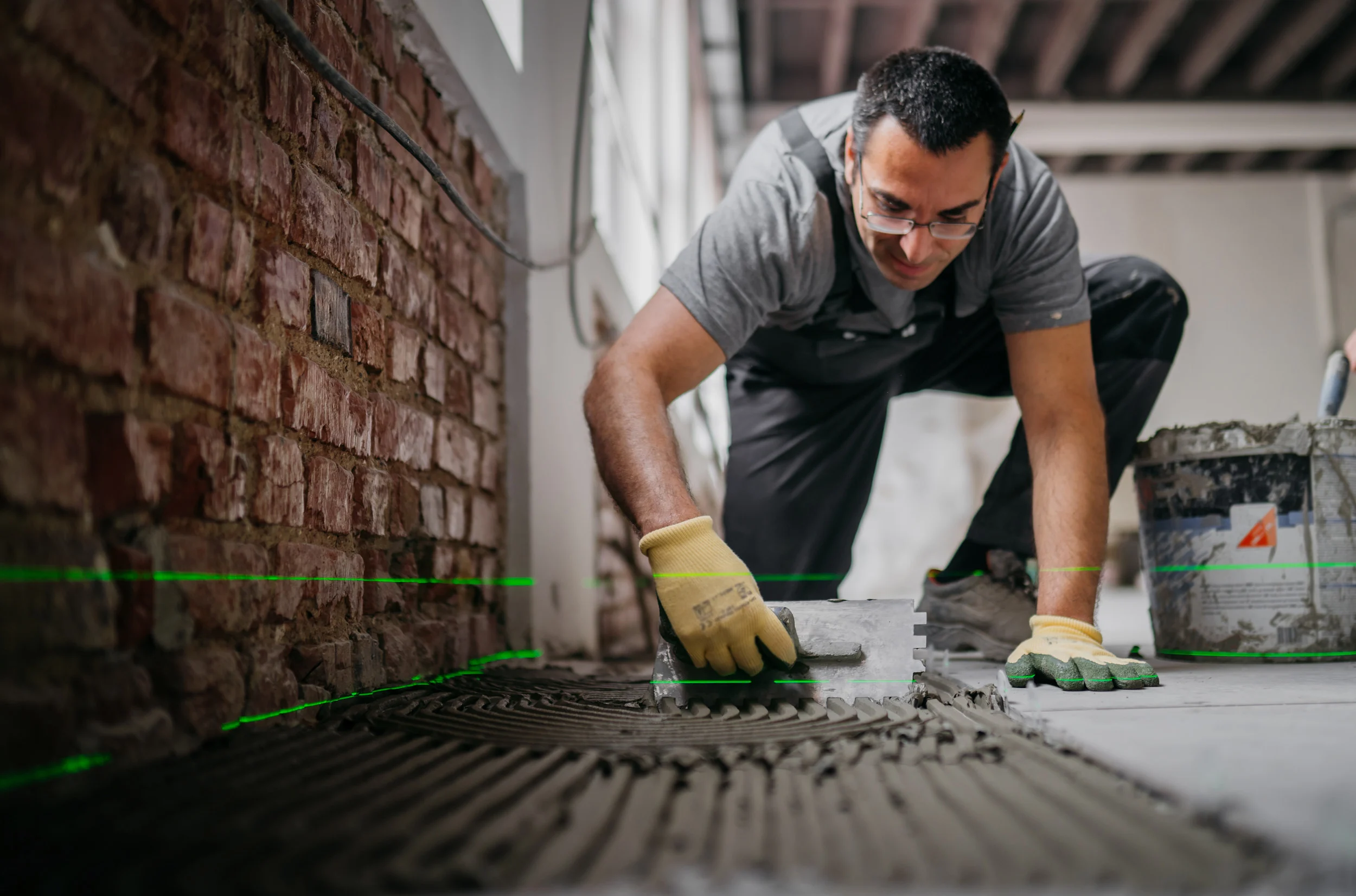 Construction worker in apron and yellow gloves laying tile against brick wall, using trowel to spread adhesive.