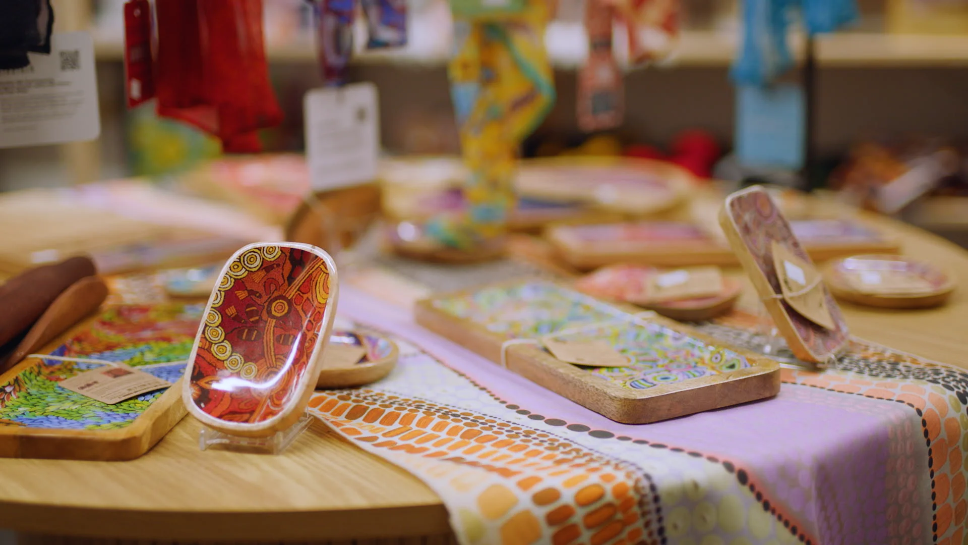 Colorful wooden trays with Aboriginal artwork displayed on a patterned tablecloth.