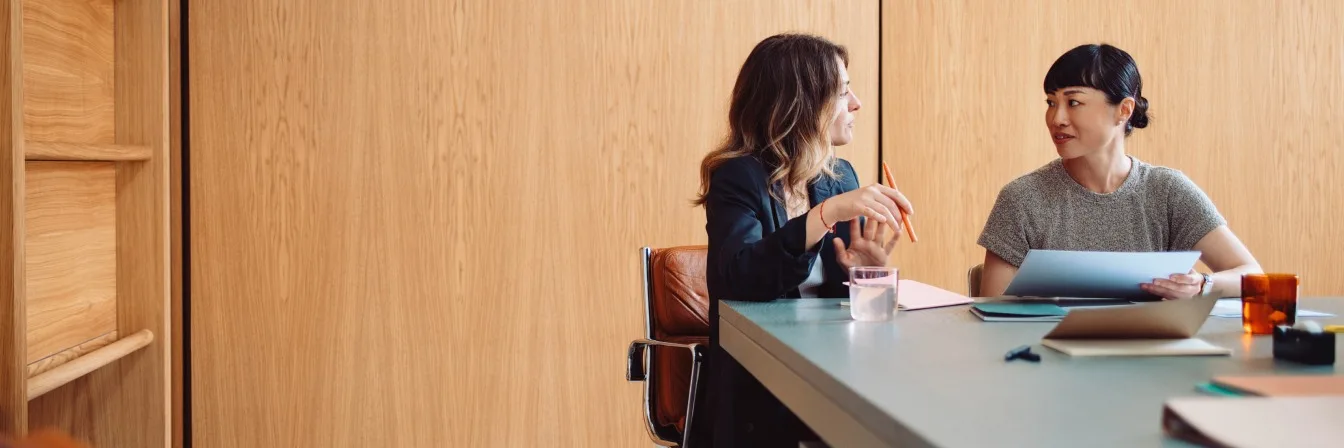 Two women in conversation at a modern office table with wooden wall panels, reviewing documents together.