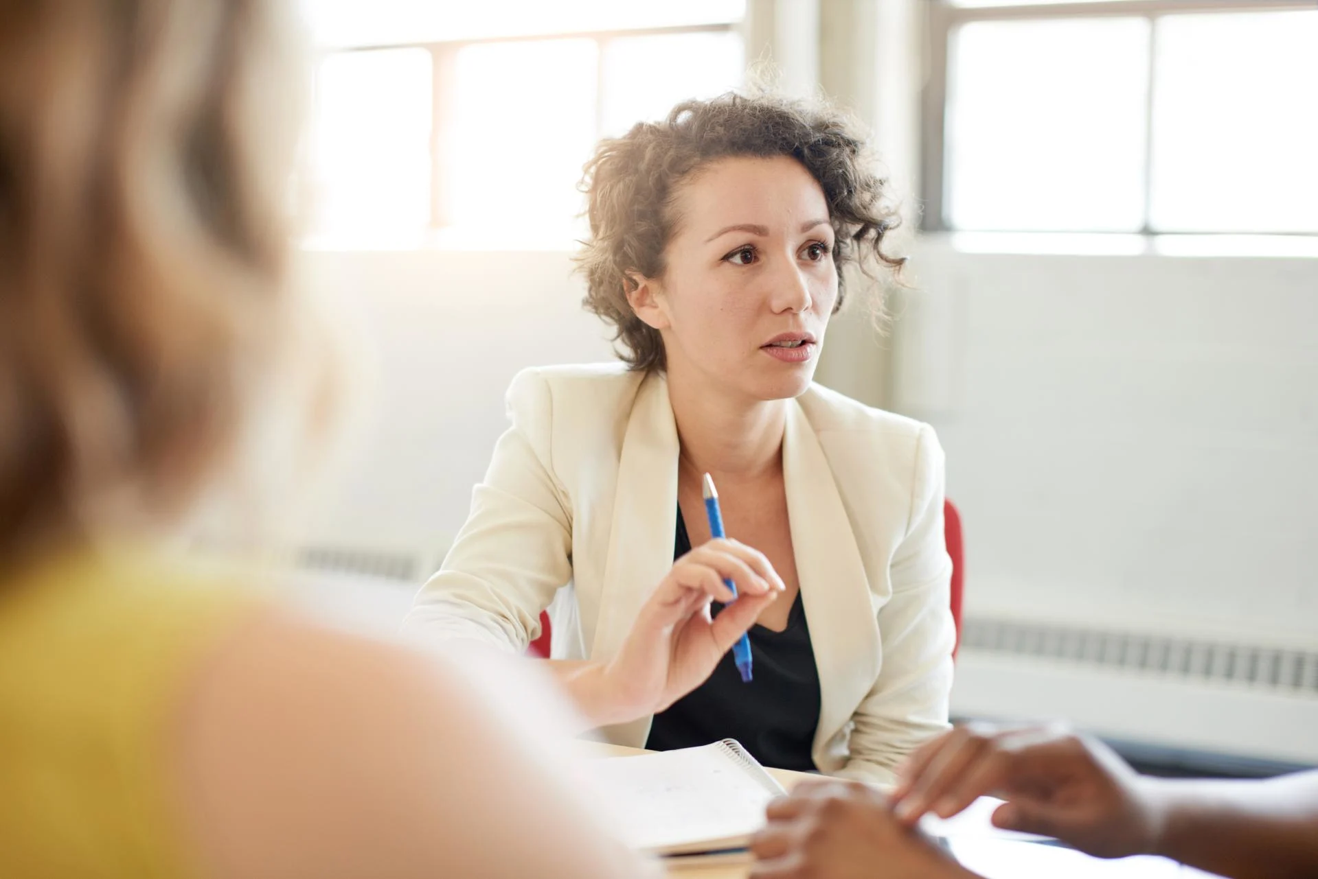 Woman with curly hair in cream blazer speaking during meeting, holding blue pen in bright office space.