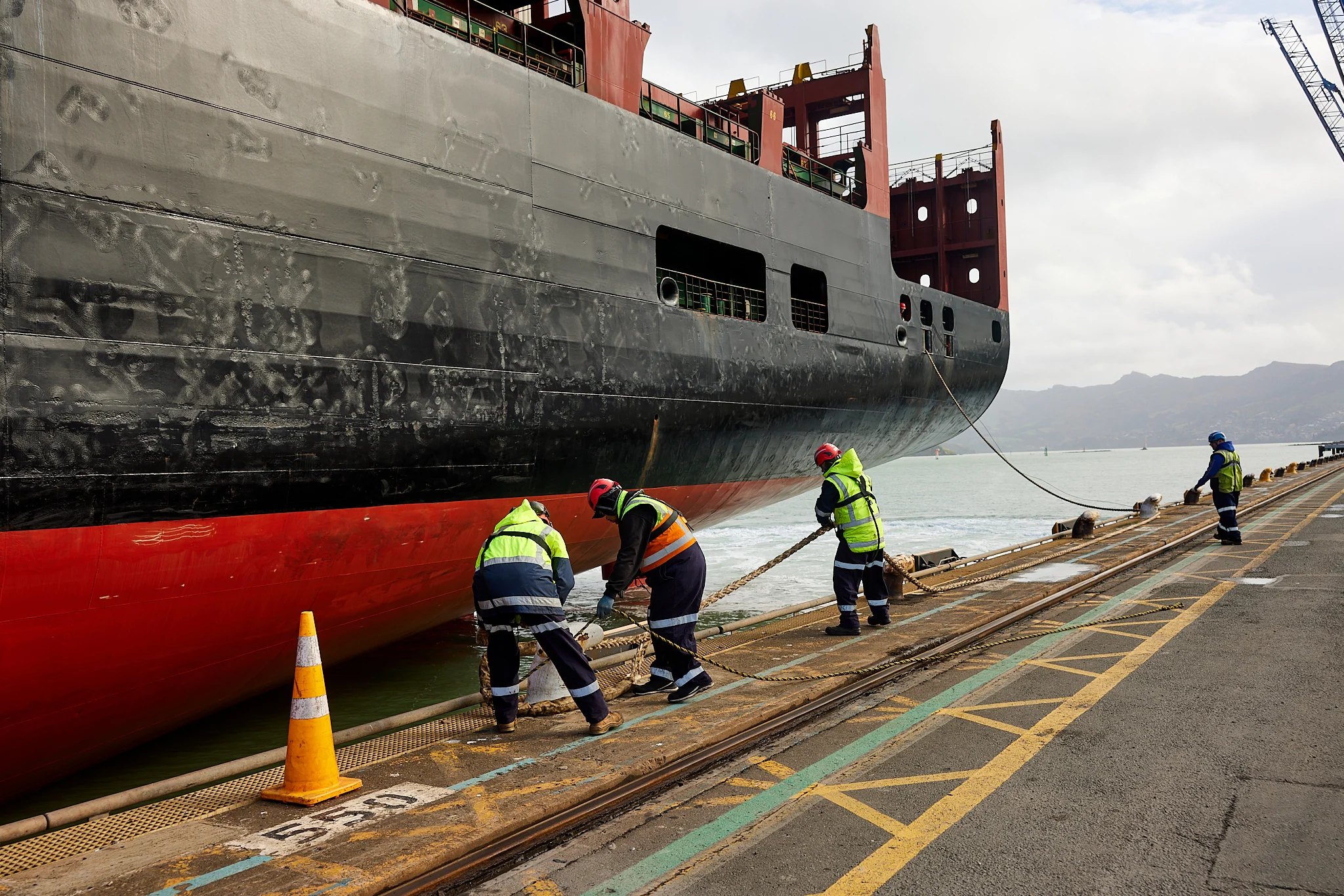 Four men in high-vis jackets and hard hats use ropes to secure a container ship that’s arrived at Lyttelton Port.