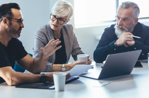 There are three people sitting at a desk in front of a laptop. One of them is holding a piece of paper and talking whilst the others look and listen.