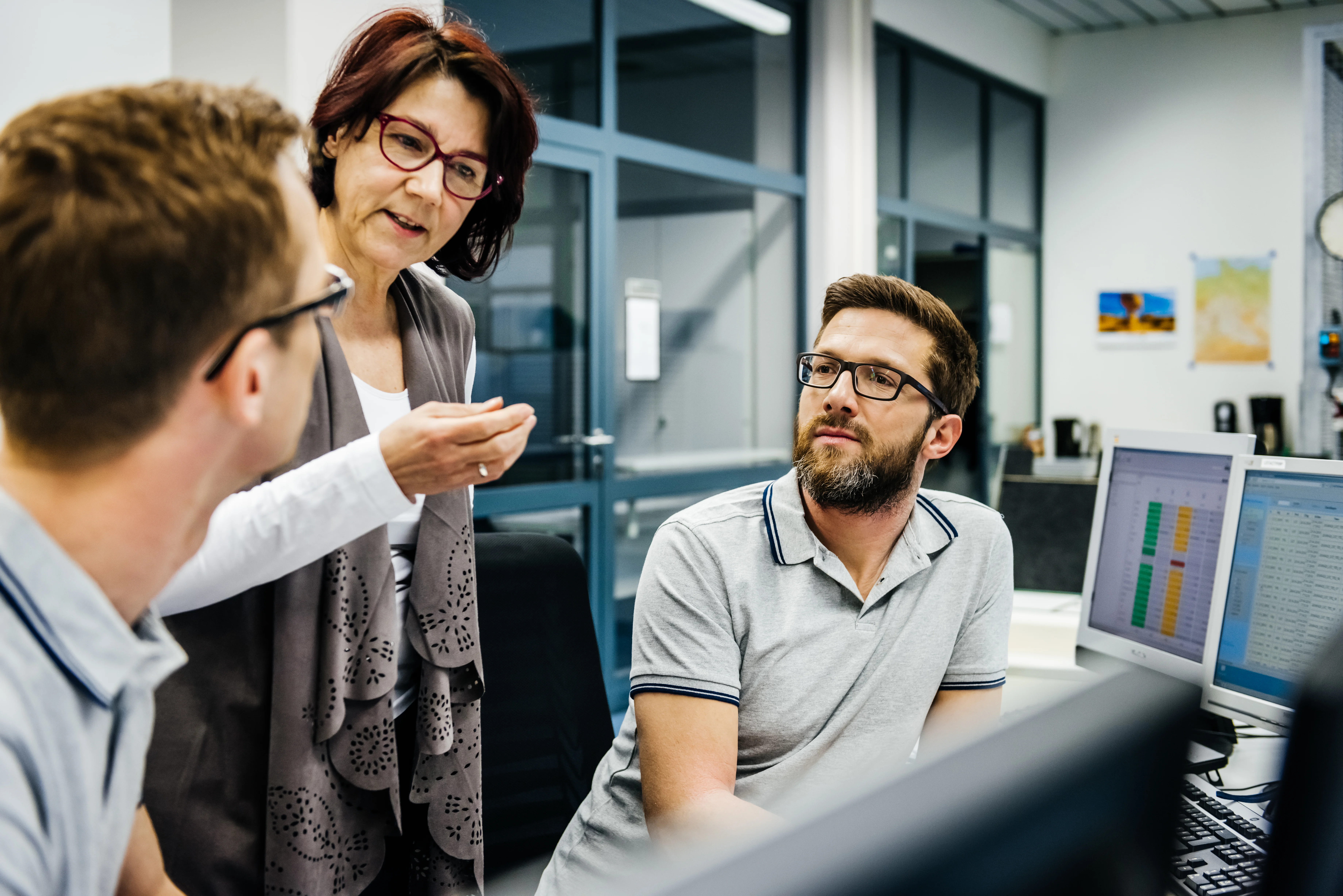 Three colleagues in discussion at an office workstation with computer monitors displaying data charts.