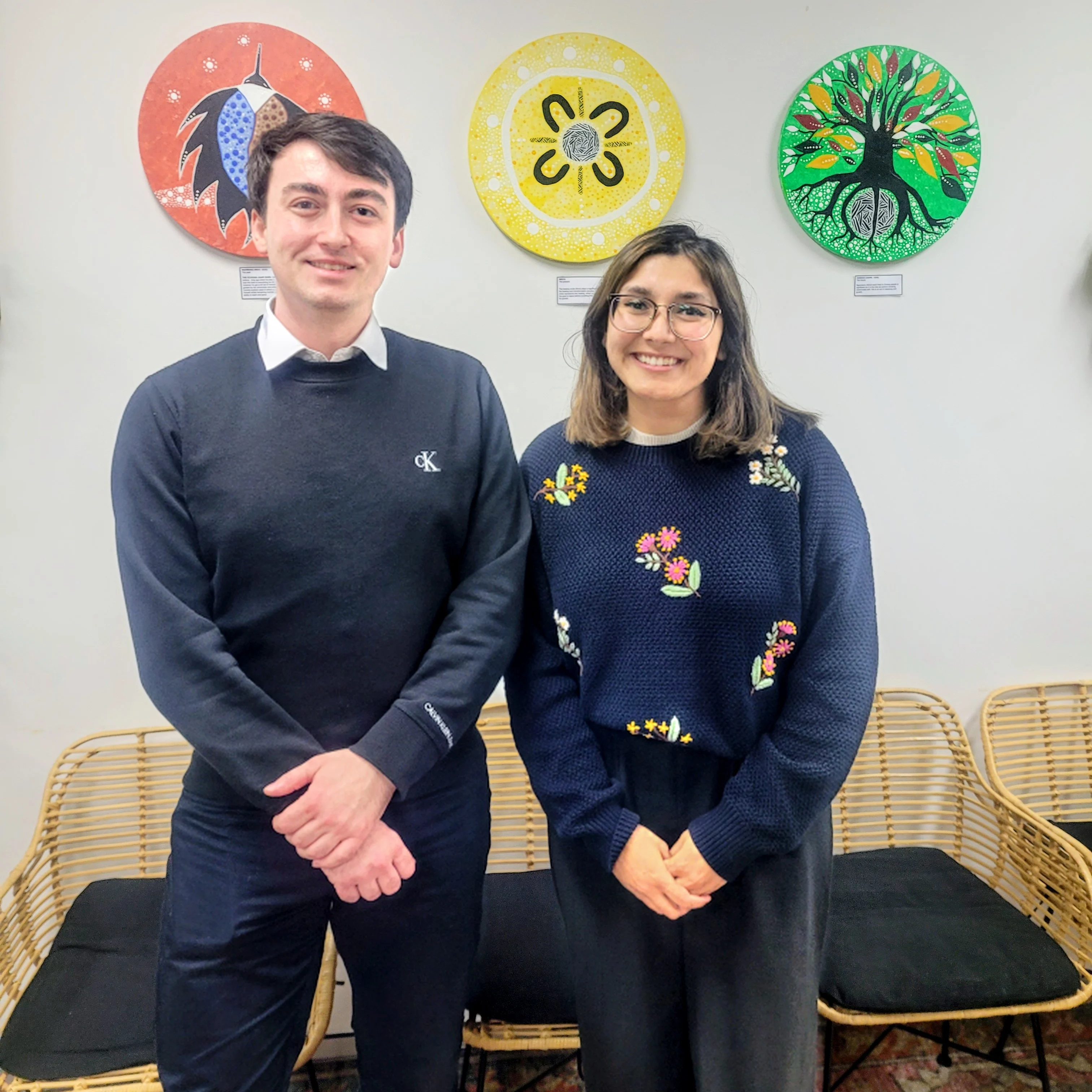 Two people in blue sweaters standing in front of colorful circular Indigenous artwork on a white wall.