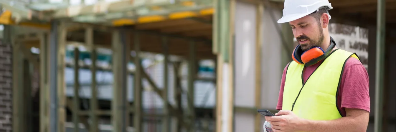 Construction worker in white hard hat and yellow safety vest checking his phone at a building site.
