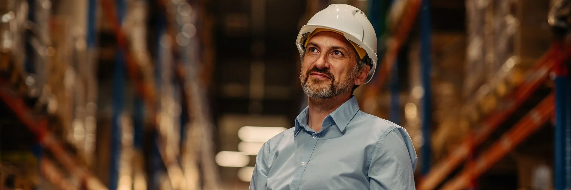 Worker in white hard hat holding tablet while looking up in warehouse with tall storage racks and ambient lighting.
