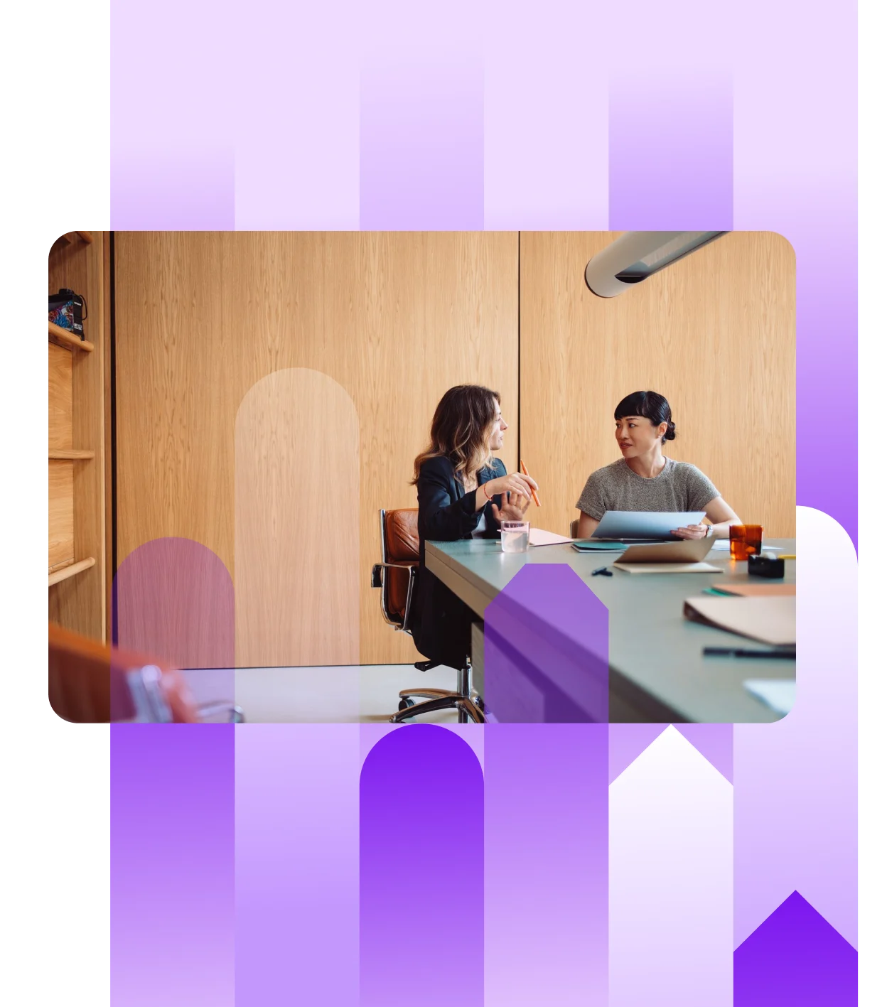 Two professional women seated at a large conference table in a modern, wood-paneled office, engaged in a conversation while reviewing documents.