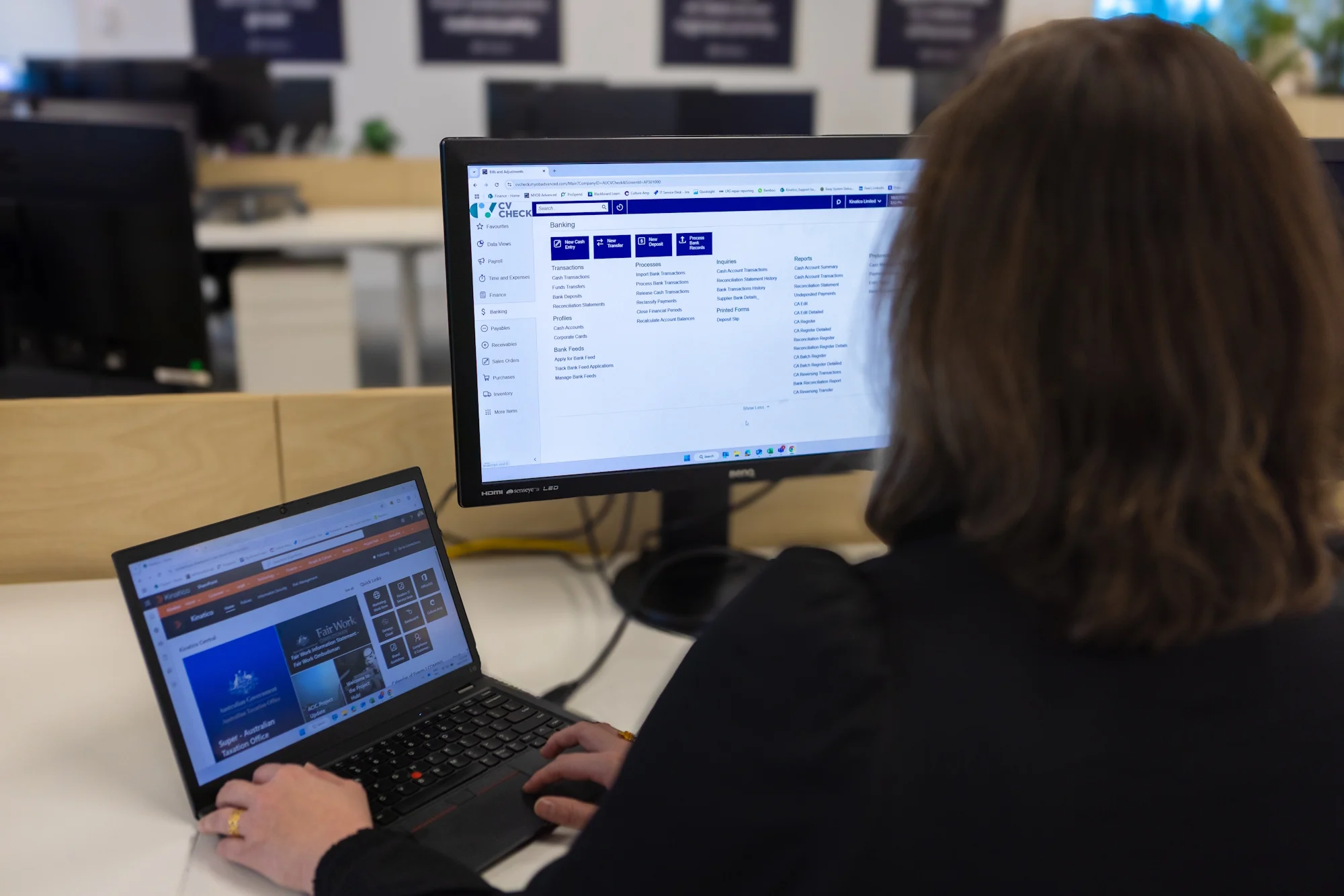 Person working at desk with laptop and monitor displaying CV Check software interface in an office environment.
