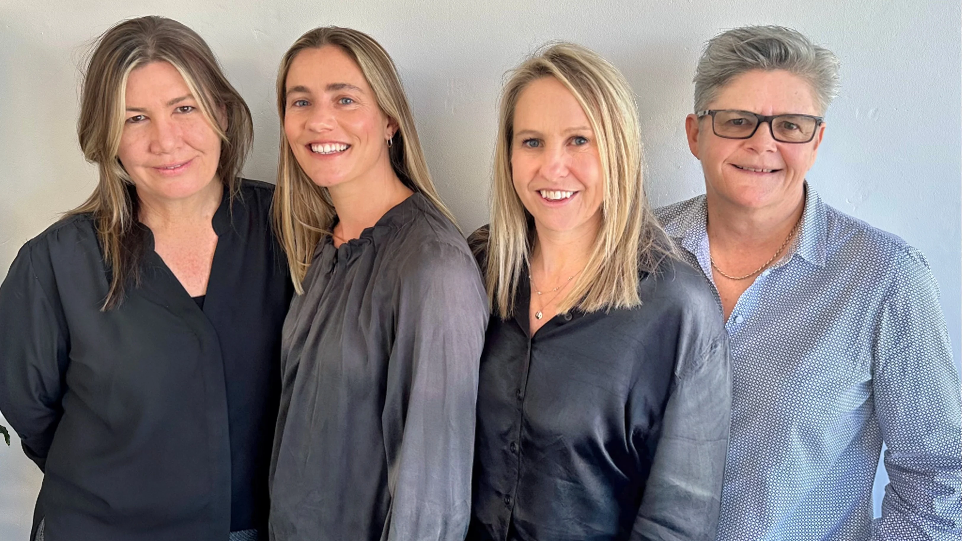 Four professionals standing side by side against a light wall, wearing dark and blue button-up shirts.