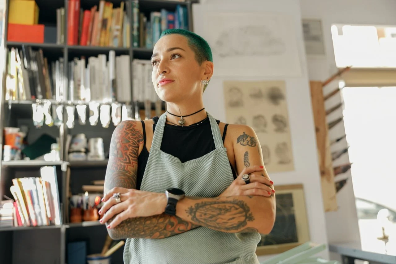 Person with green hair and tattoos wearing striped apron in creative workspace with bookshelves in background.