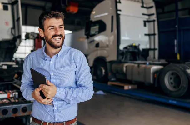 Professional logistics worker smiles with tablet under their arm. There are empty trucks in the background.