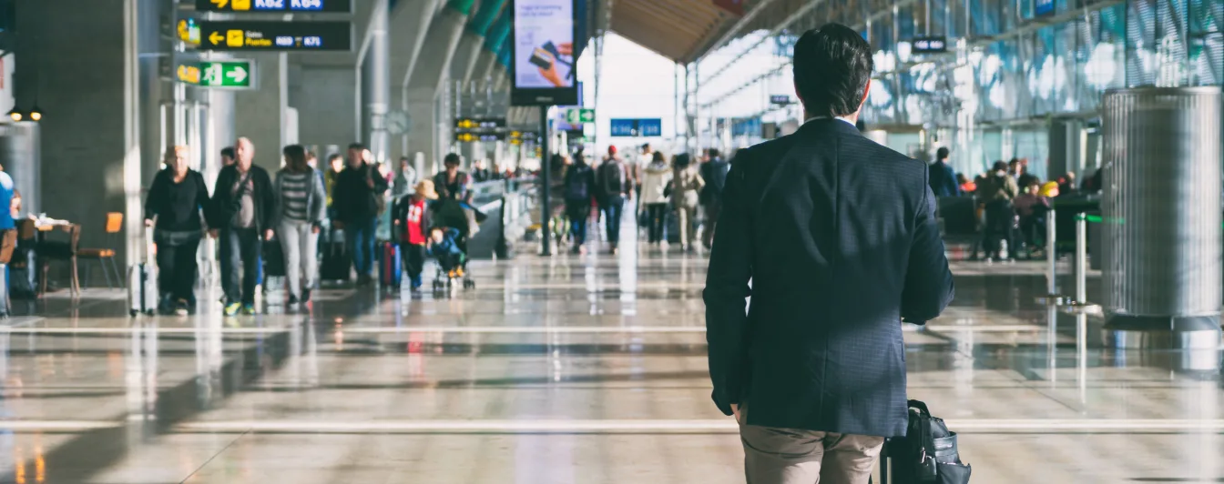Person in suit walking through busy airport terminal with directional signs and travelers visible in background.