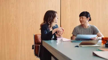 Two women sitting at a table having a conversation in a meeting room with wooden wall panels.