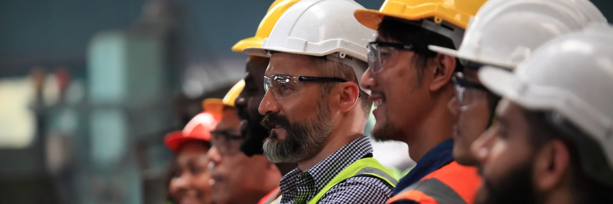 A diverse group of workers wearing hard hats and safety vests in yellow, orange, and white stand together in an industrial setting.