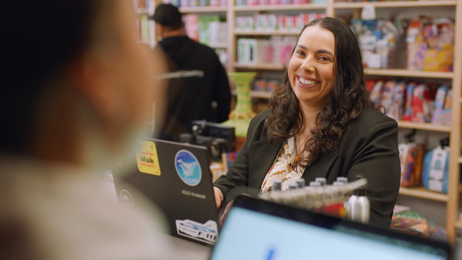 Woman with curly hair in green blazer smiling at customer in retail store with colorful shelves in background.