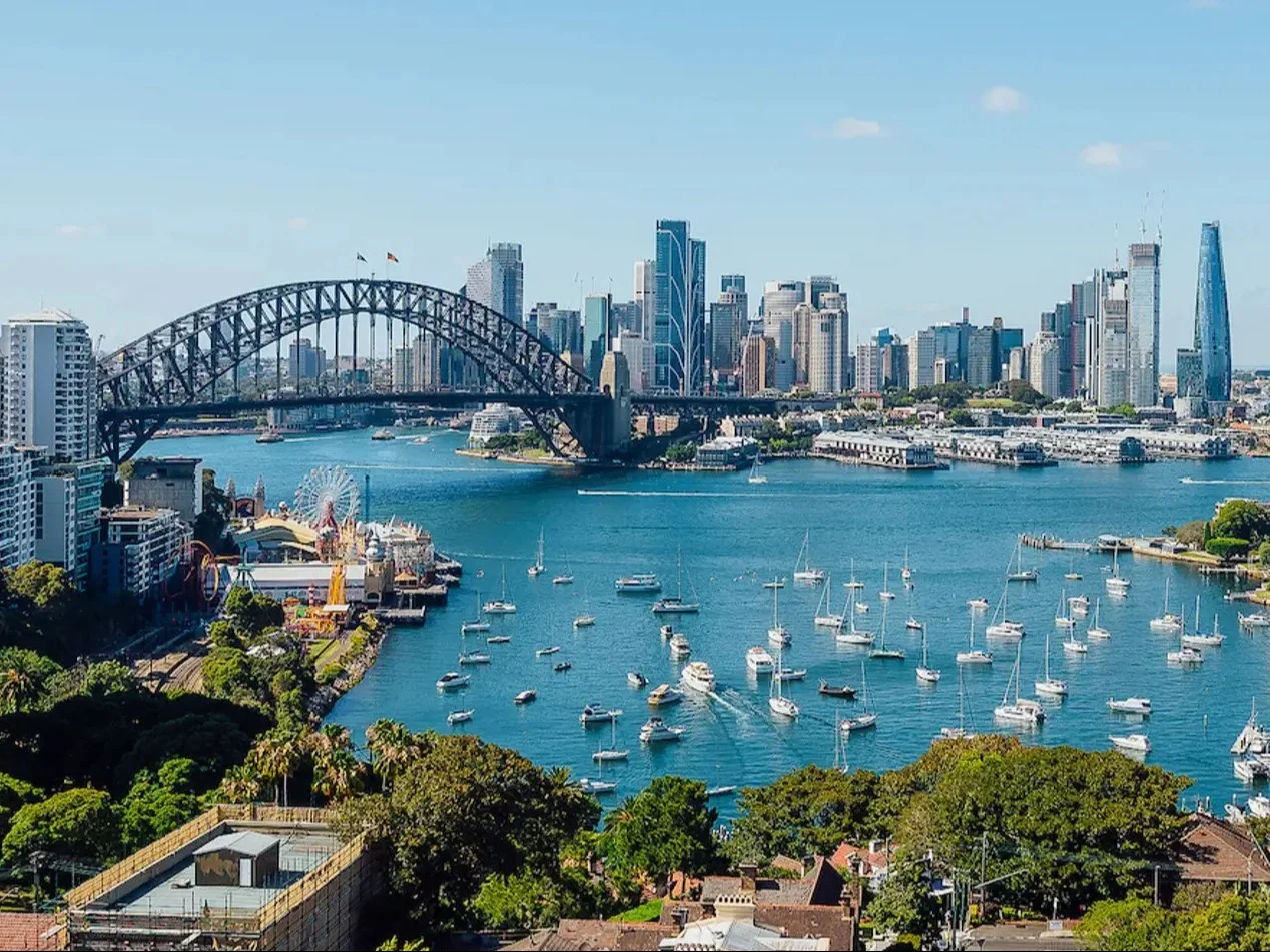 Sydney Harbour panorama with iconic bridge, city skyline, and boats dotting the blue water under clear skies.