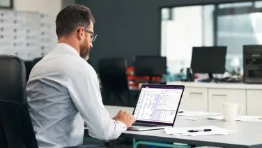 Person in white shirt working on laptop in modern office with data spreadsheet visible on screen