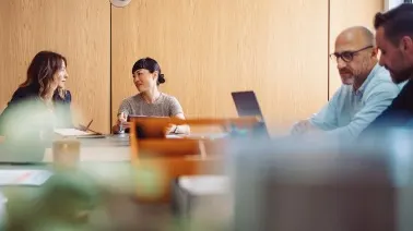 People in a business meeting around a wooden conference table with laptops, engaged in discussion.