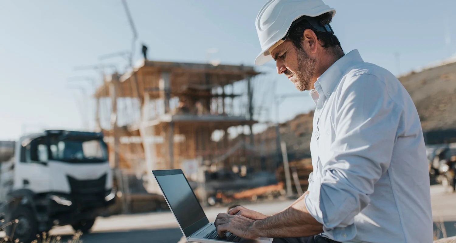 Construction professional in white hard hat working on laptop at building site with framework structure in background.