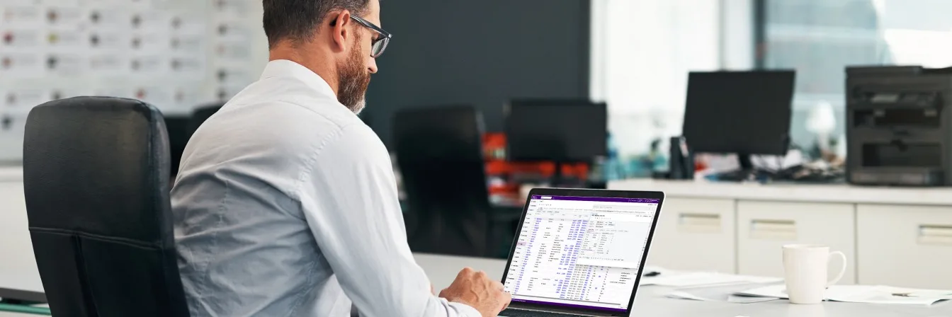 Person in white shirt working on laptop in modern office with data spreadsheet visible and coffee mug nearby.