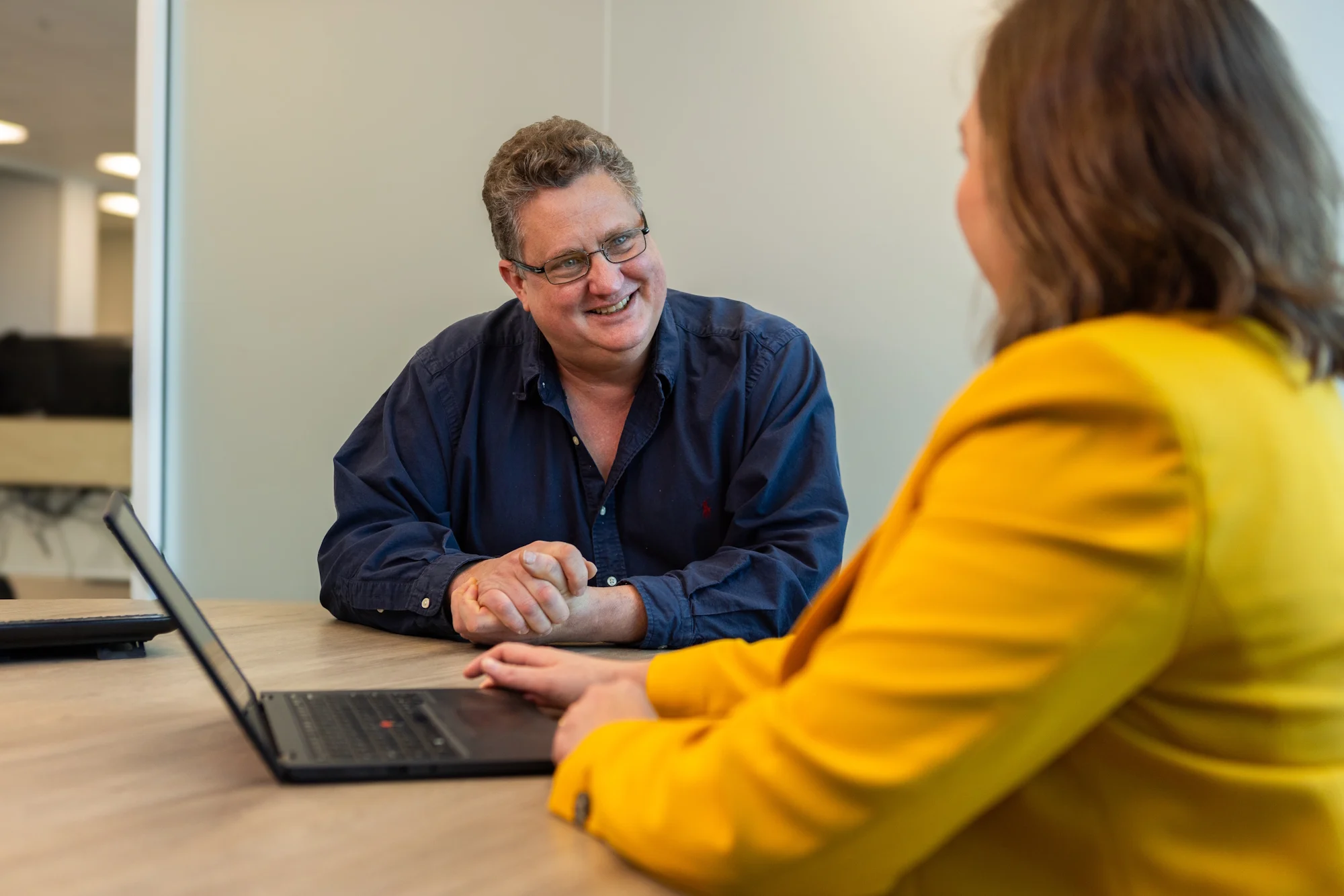 Two people in conversation at a wooden table with a laptop; person in blue shirt smiling at person wearing bright yellow jacket.