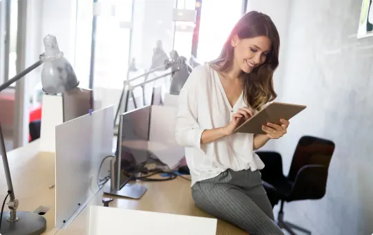 A woman with long brown hair sitting on an office desk uses a tablet while smiling. 