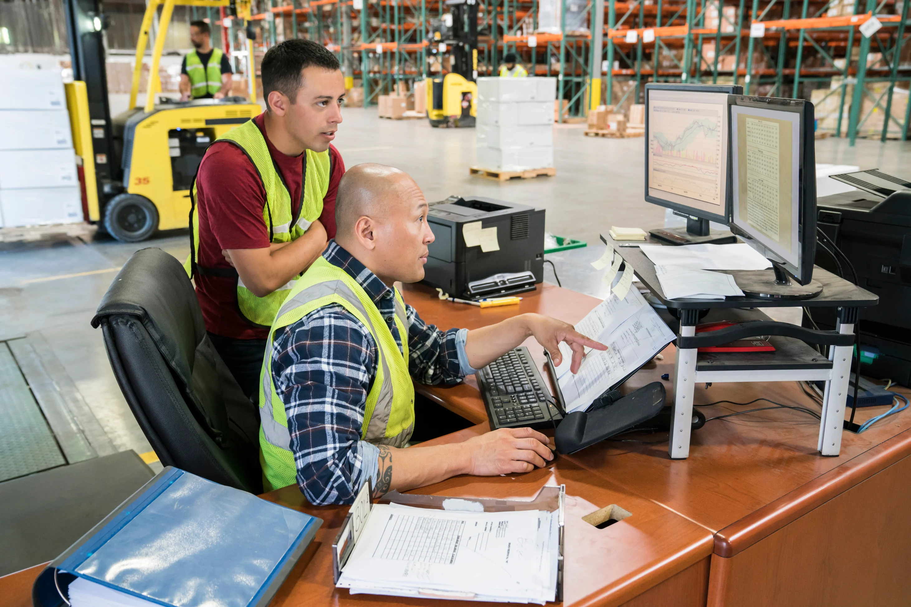 Warehouse workers in safety vests reviewing inventory data on computer monitors with forklift and storage racks in background.