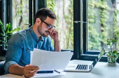 Financial services professional worker sits at desk analysing data on laptop and printouts. 