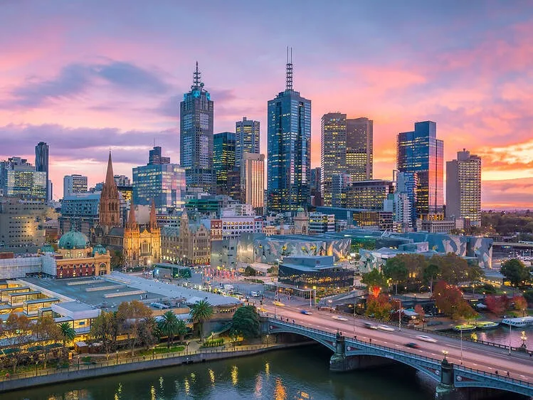 Melbourne skyline at sunset with illuminated skyscrapers, historic buildings, and Yarra River in the foreground.