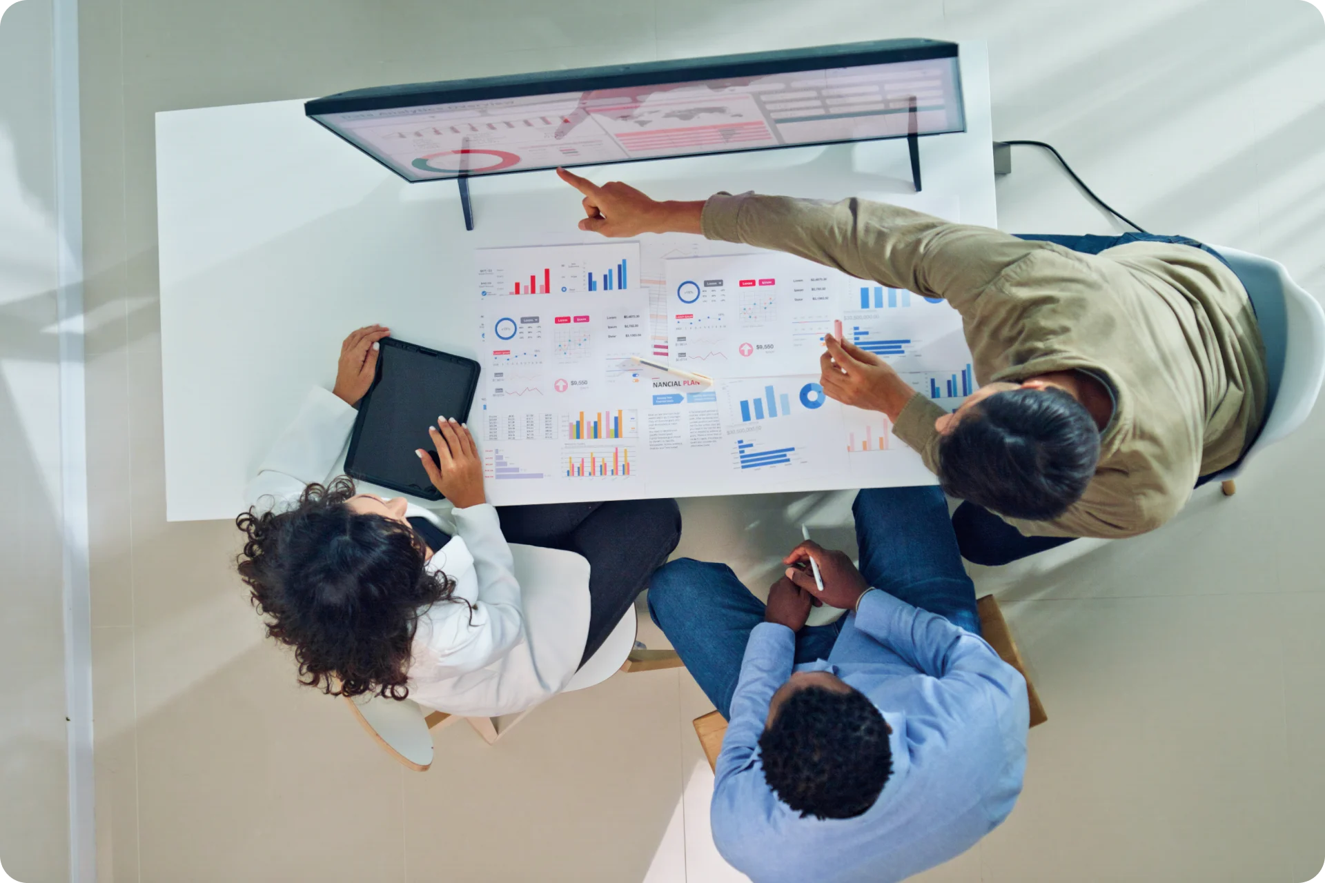 Overhead view of three professionals analyzing colorful data charts and graphs on a desk, with one person pointing at the display.