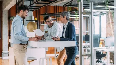 Three professionals reviewing architectural plans together at a standing desk in a modern office with exposed brick walls.